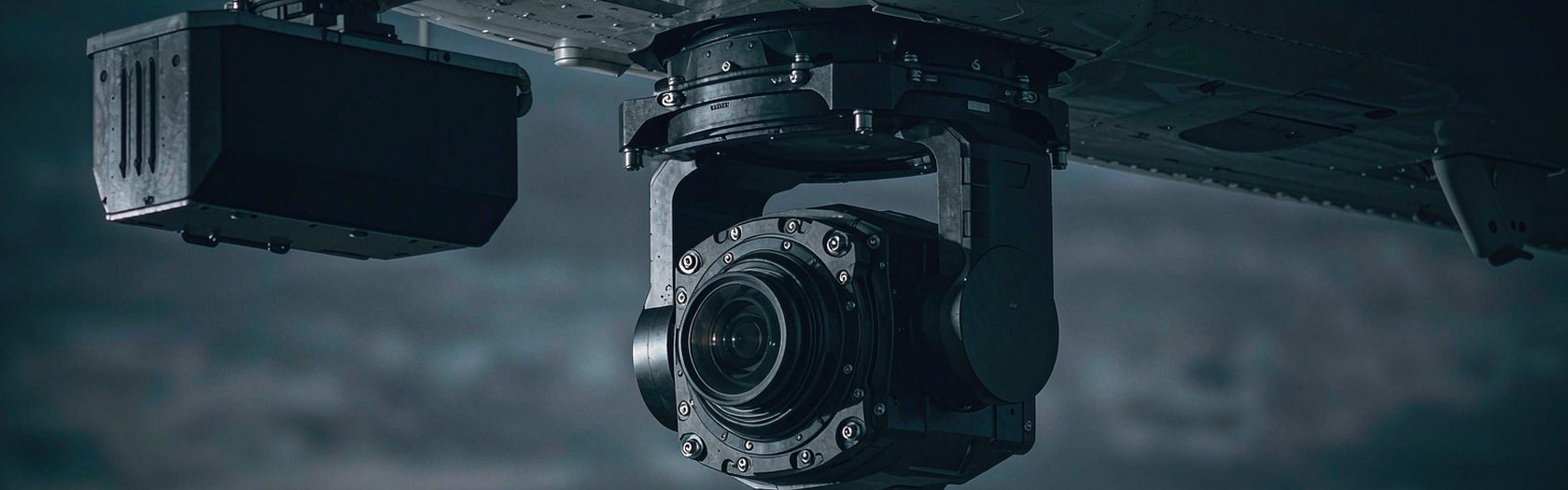 An airborne electro-optical gimbal turret under the fuselage of an unmarked survey aircraft at dusk.